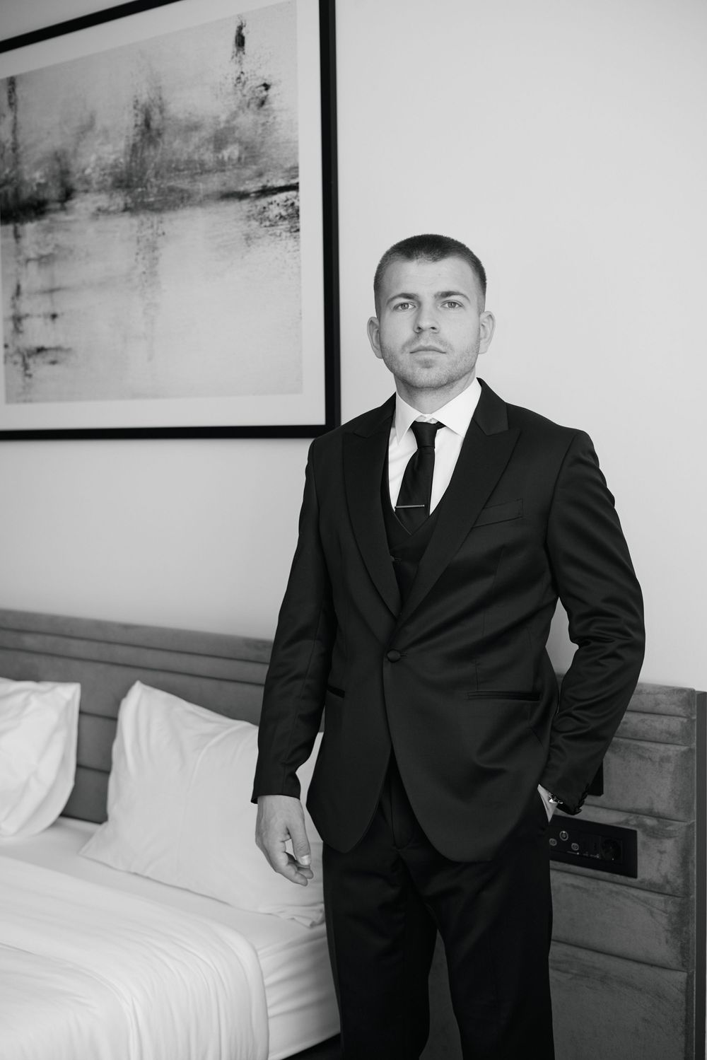 Elegant groom in black suit and tie posing in hotel room before wedding ceremony.
