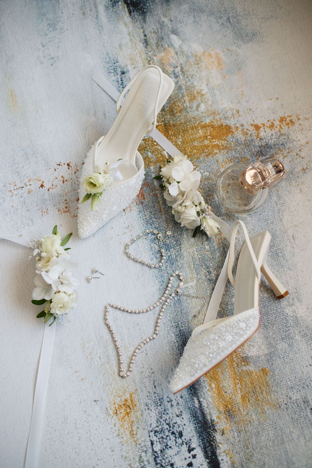 Bridal accessories flat lay with white lace heels, pearl necklace, flowers, and perfume bottle.