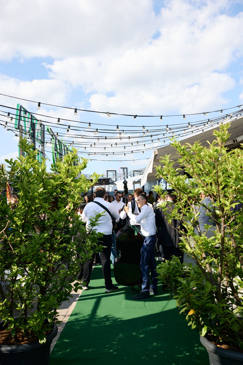 Wedding guests walk through lush green garden path with string lights overhead at outdoor venue.