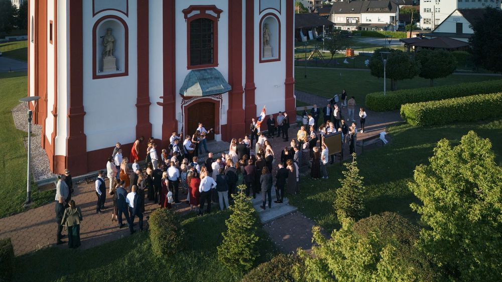 Aerial view of a crowd gathering outside a red and white church surrounded by green trees and lawn.