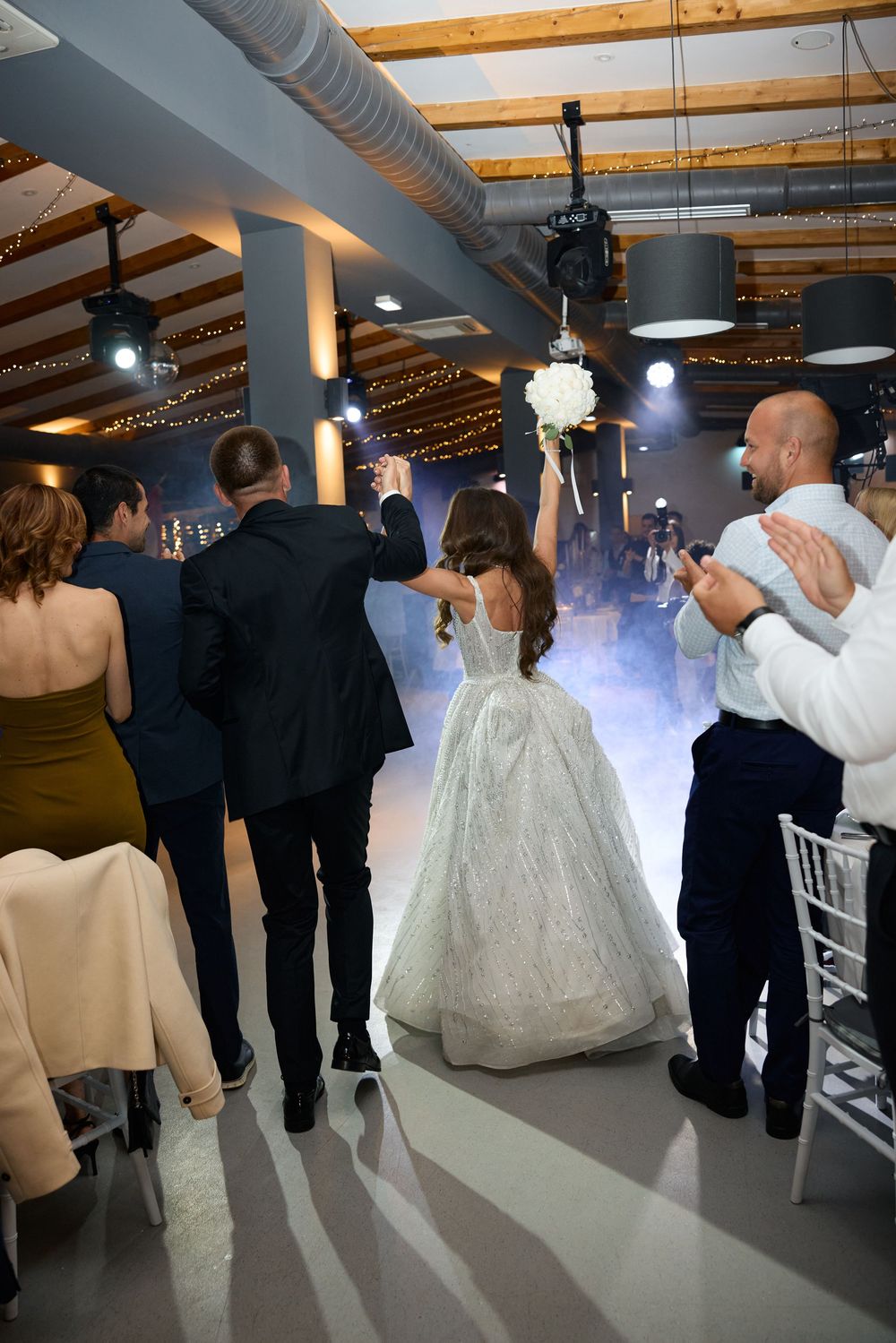 Bride and groom make grand entrance at wedding reception, bride holding bouquet aloft amid dry ice fog.