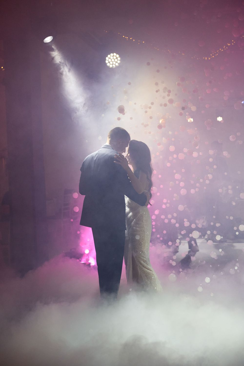 Newlyweds sharing first dance surrounded by romantic fog and bokeh lights at wedding reception.