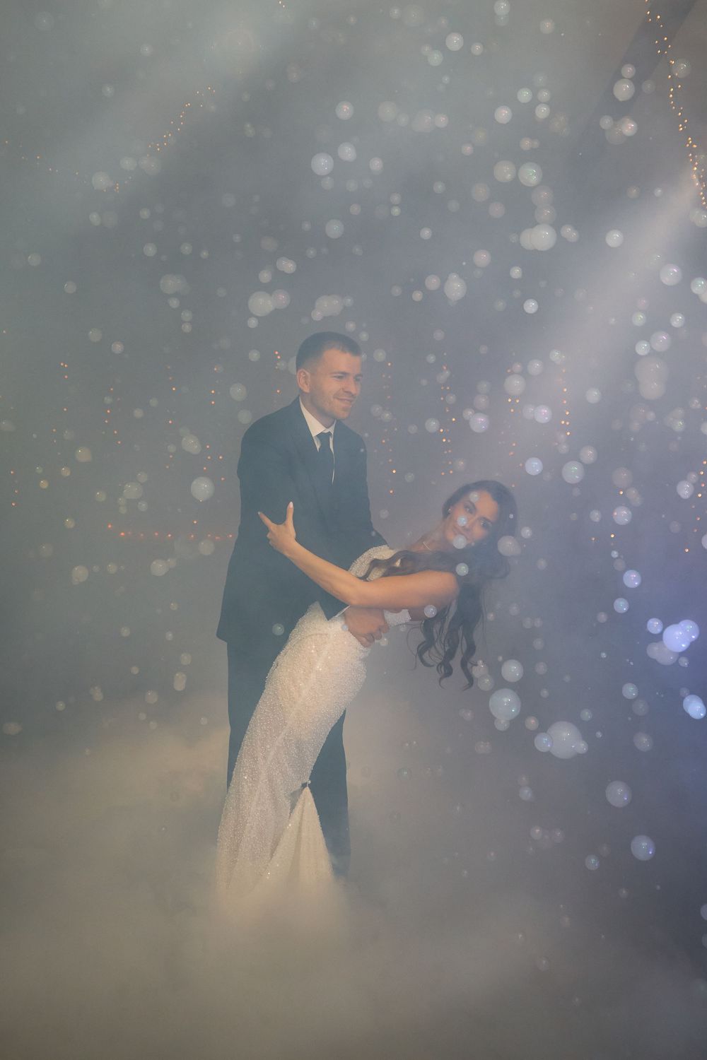 Bride and groom dancing romantically surrounded by fog and bokeh lights at wedding.