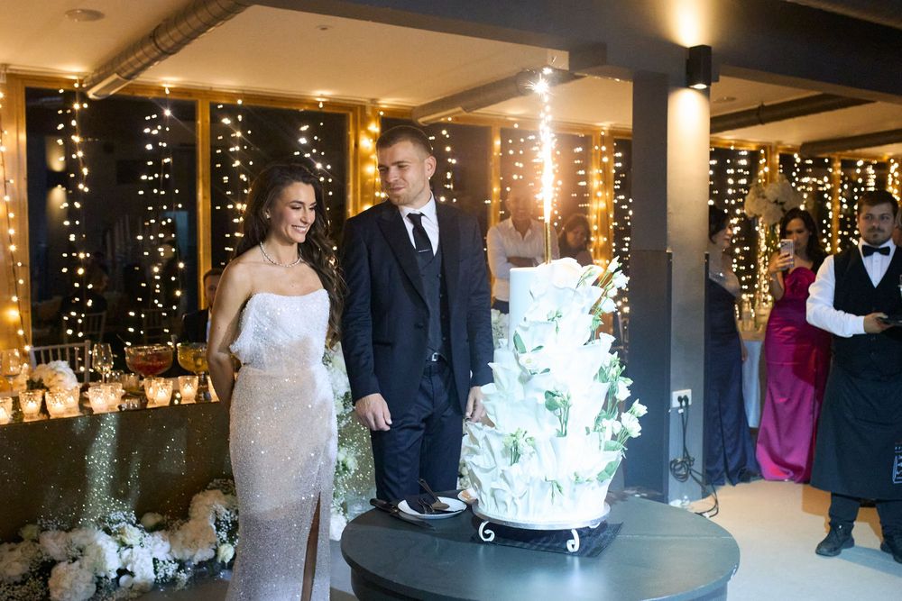 Elegant couple cutting a white floral wedding cake in a warmly lit reception venue with fairy lights.