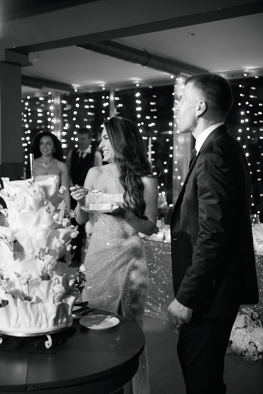 Black and white photo of bride and groom cutting wedding cake surrounded by fairy lights.