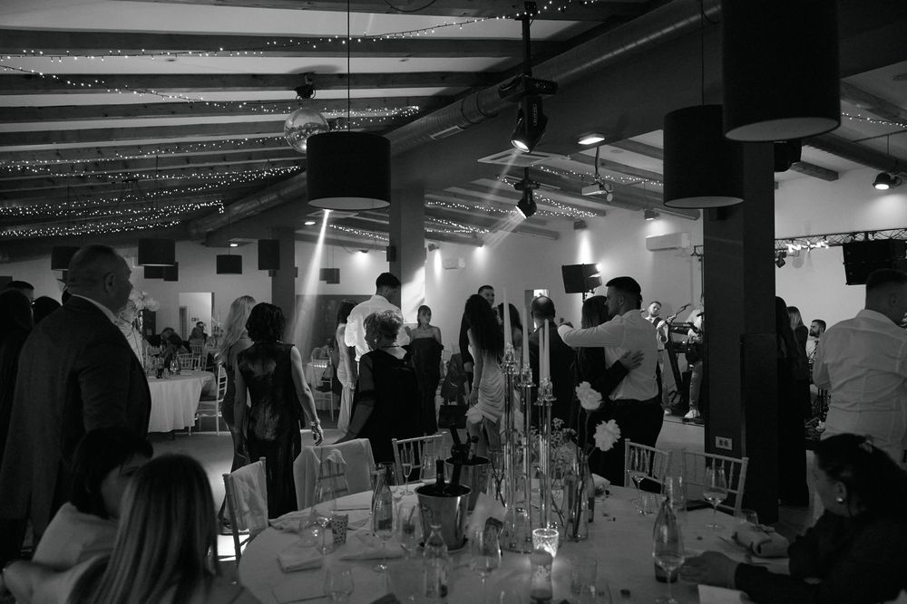 Black and white wide-angle shot of wedding guests dancing in a decorated reception hall with fairy lights.