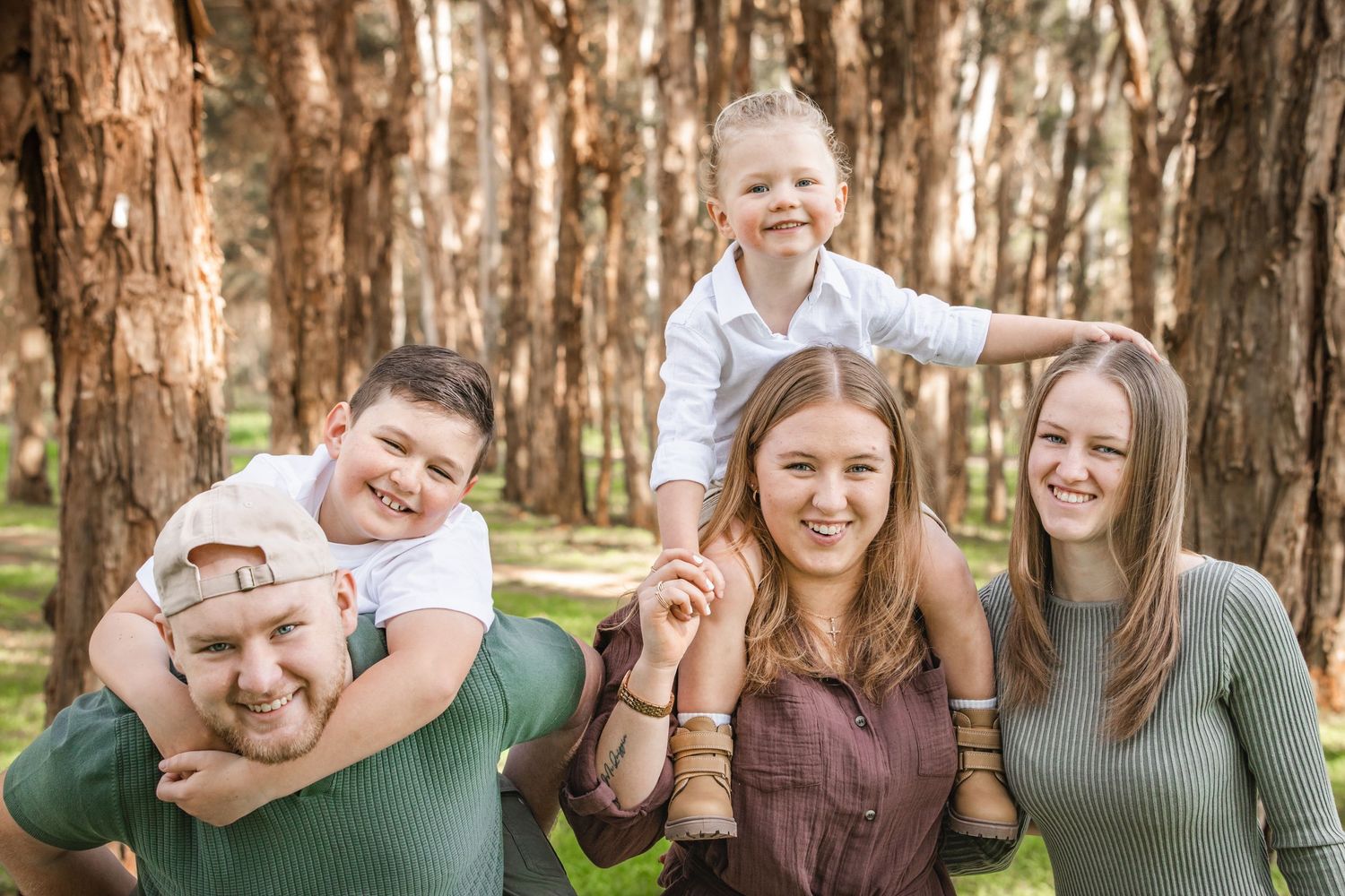 Family of five smiling outdoors among tall trees, with children on shoulders and holding hands in a sunny forest in the Hills District.
