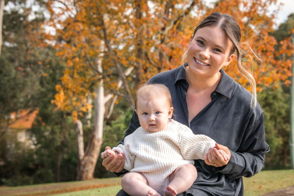 Parent and infant smile together outdoors on a sunny fall day with orange trees in the background.