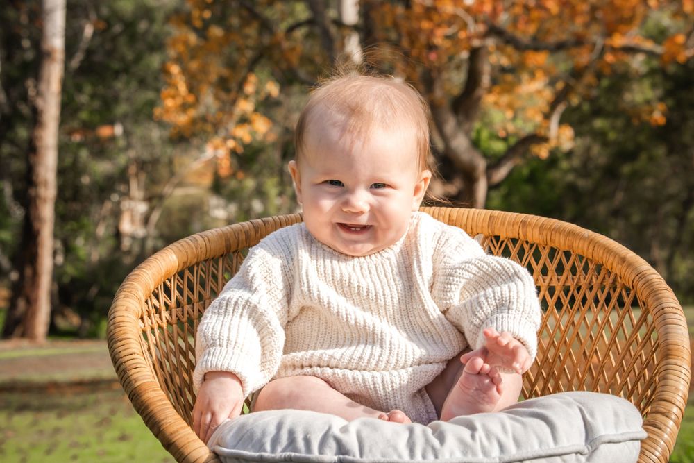 Baby in white sweater sitting in wicker chair outdoors during autumn season.