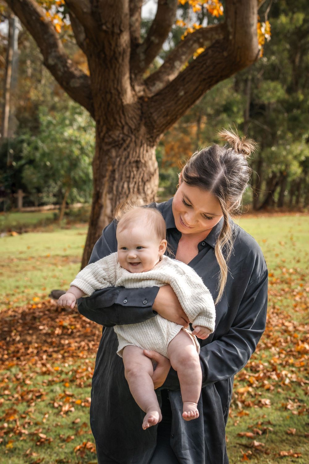 Autumn scene in park with baby wearing cream sweater enjoying outdoor moment under large tree with fallen leaves in Castle Hill.
