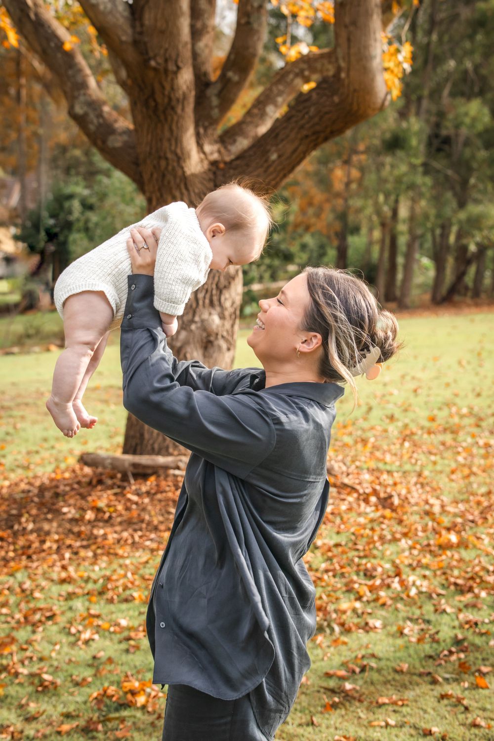 A joyful moment between parent and baby in an autumn park with fallen leaves beneath a golden-hued tree.