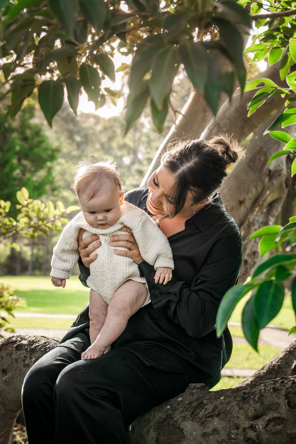 A mother sits on a tree branch in a lush park, lovingly holding her baby dressed in a cream knit outfit at Castle Hill Heritage Park.