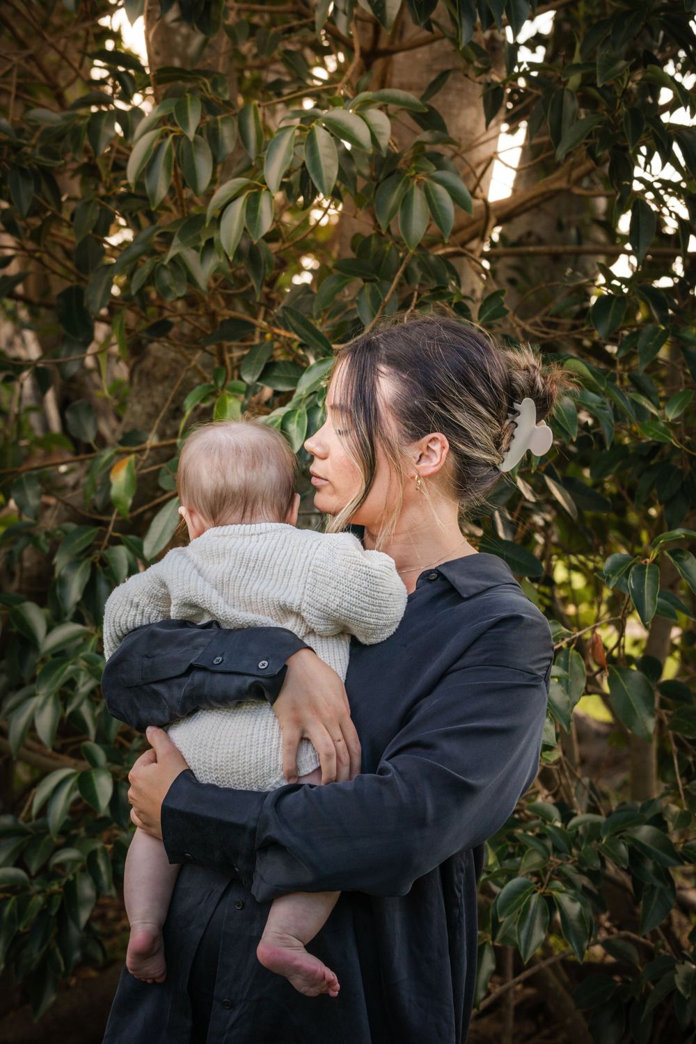 A parent in a navy sweater holds a baby wearing a beige knit sweater against a backdrop of green foliage.