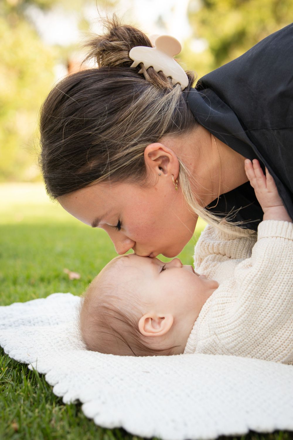Mother and baby photo sharing a tender moment while laying on a white blanket in a sunny outdoor setting.