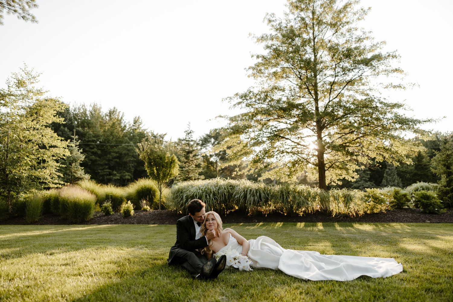 Bride and groom sitting together on lush green lawn surrounded by trees and garden landscaping in golden hour light.
