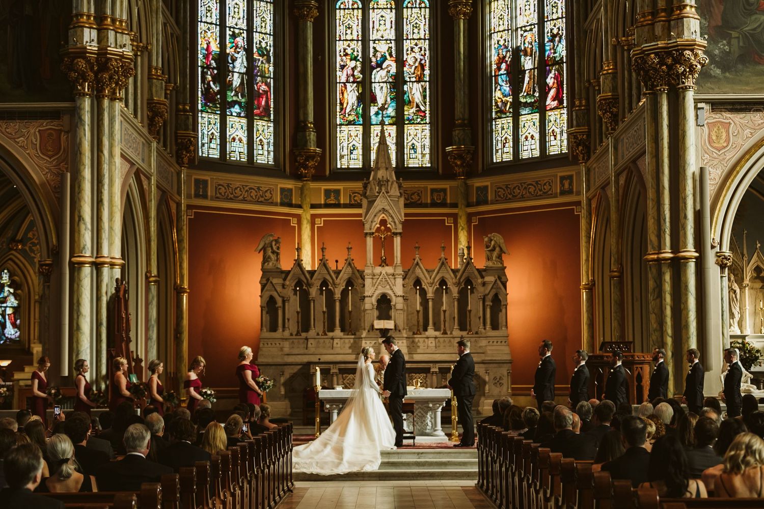 Bride and groom exchange vows at ornate church altar with stunning stained glass windows and seated wedding guests.