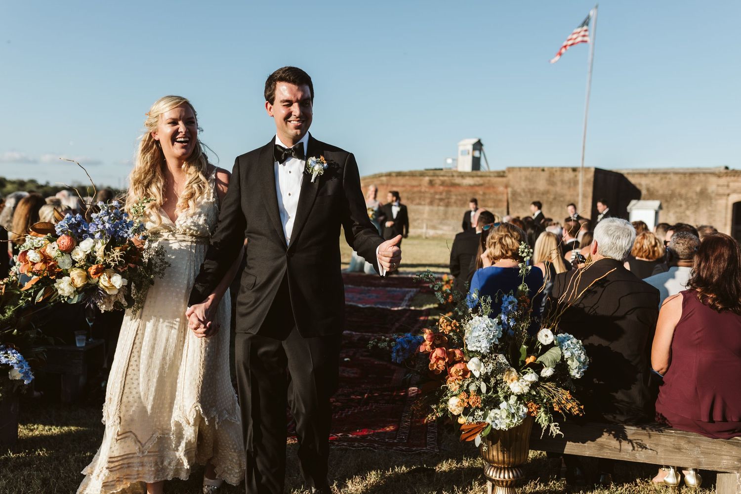Bride and groom walk down the aisle at an outdoor wedding ceremony with colorful floral arrangements.