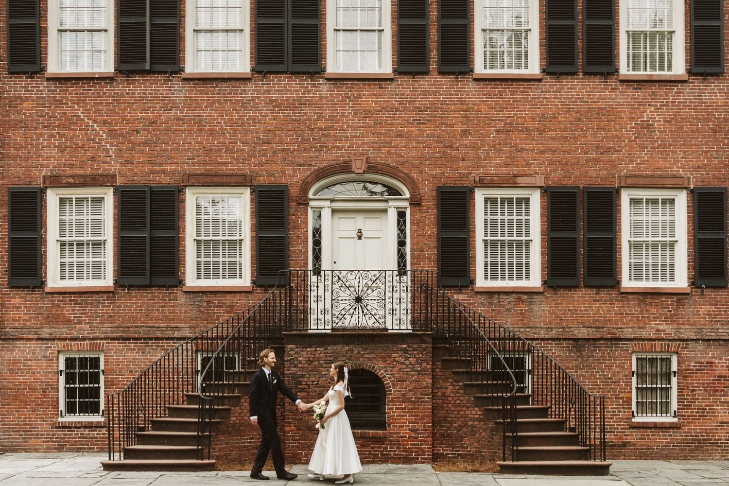 Bride and groom hold hands outside a historic red brick building with black shutters and iron staircases.