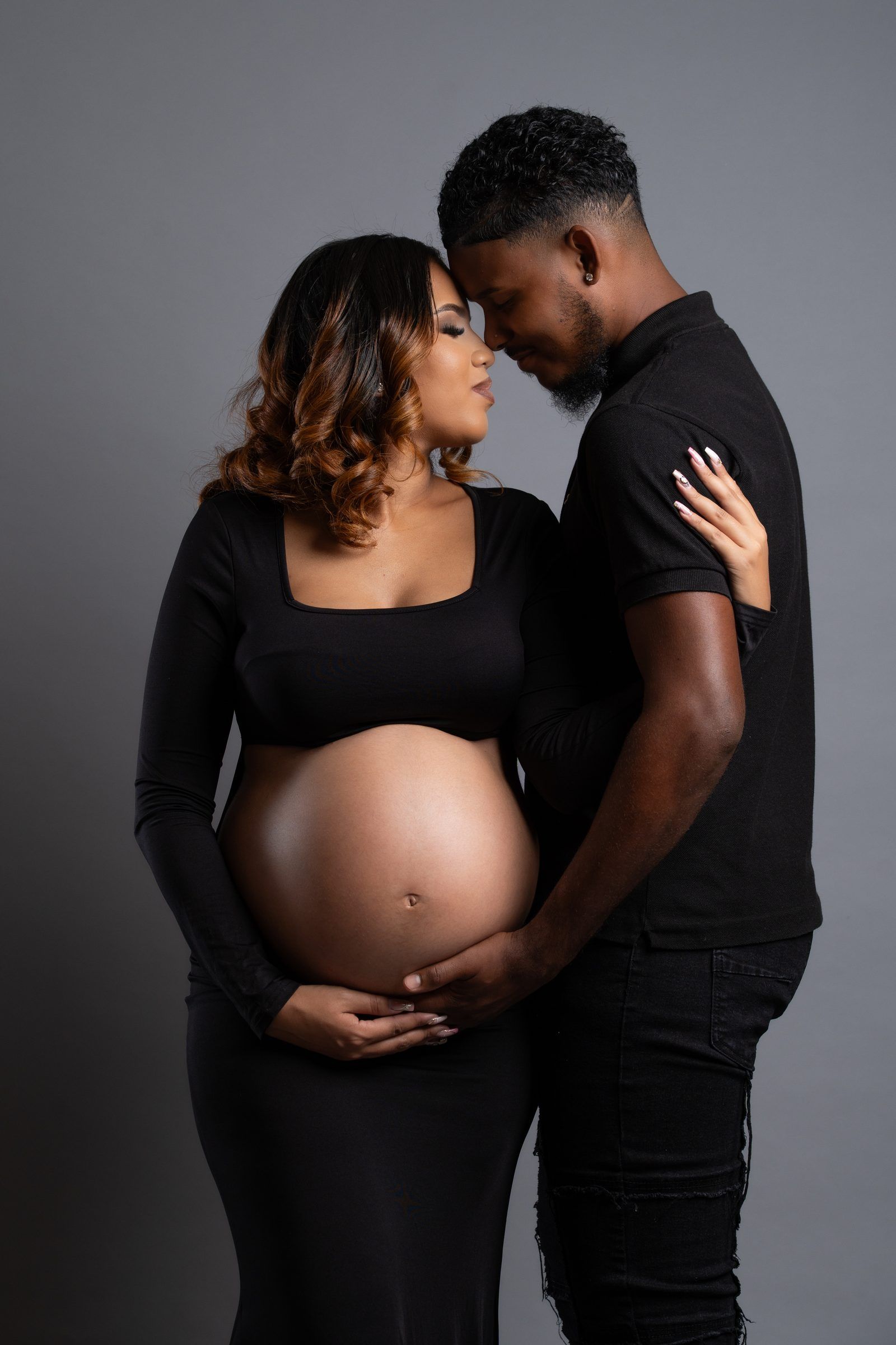 A pregnant Black couple in matching black outfits share an intimate moment, foreheads touching, cradling her baby bump.