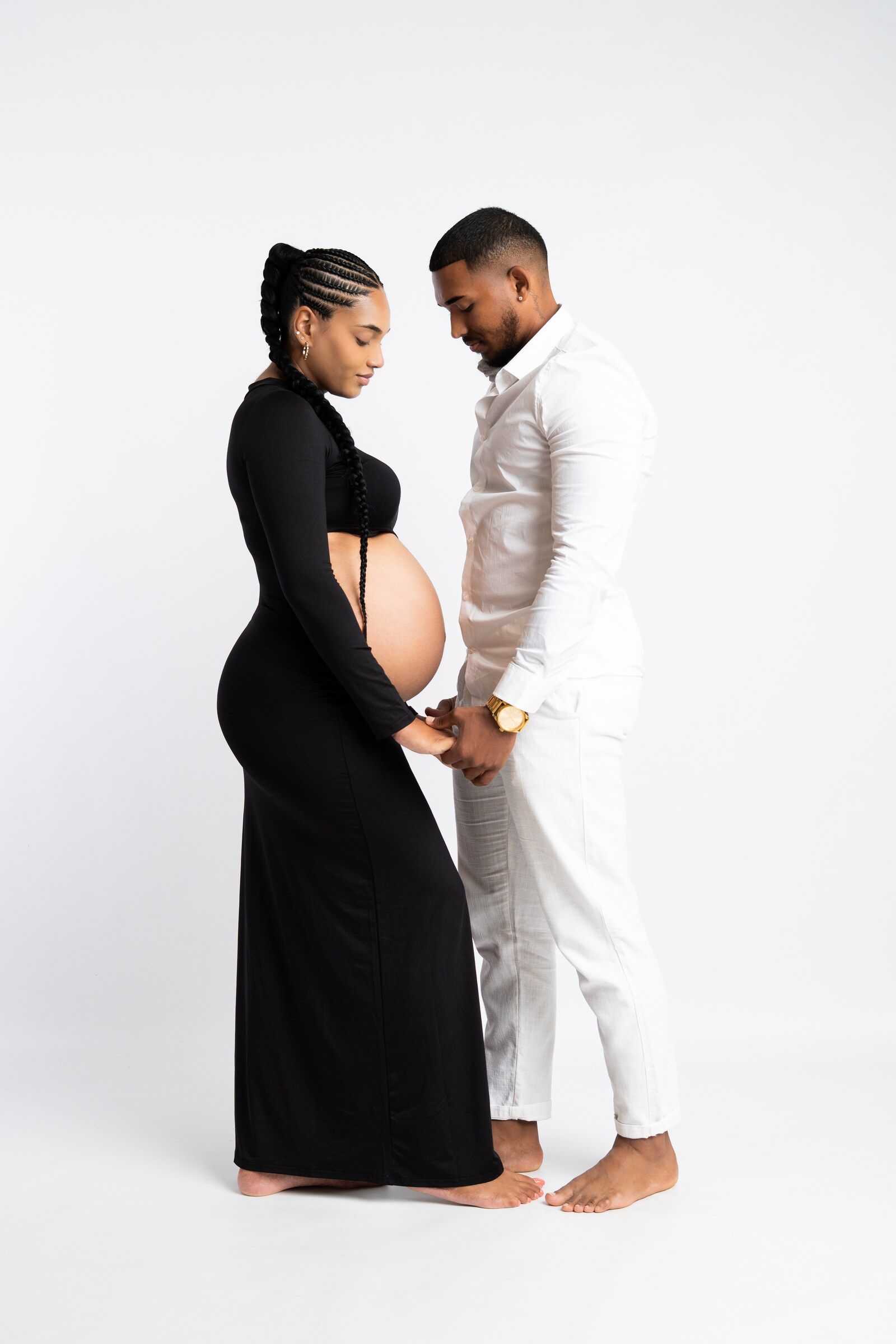 A pregnant couple in black and white outfits holding hands, looking down at her baby bump in a studio.