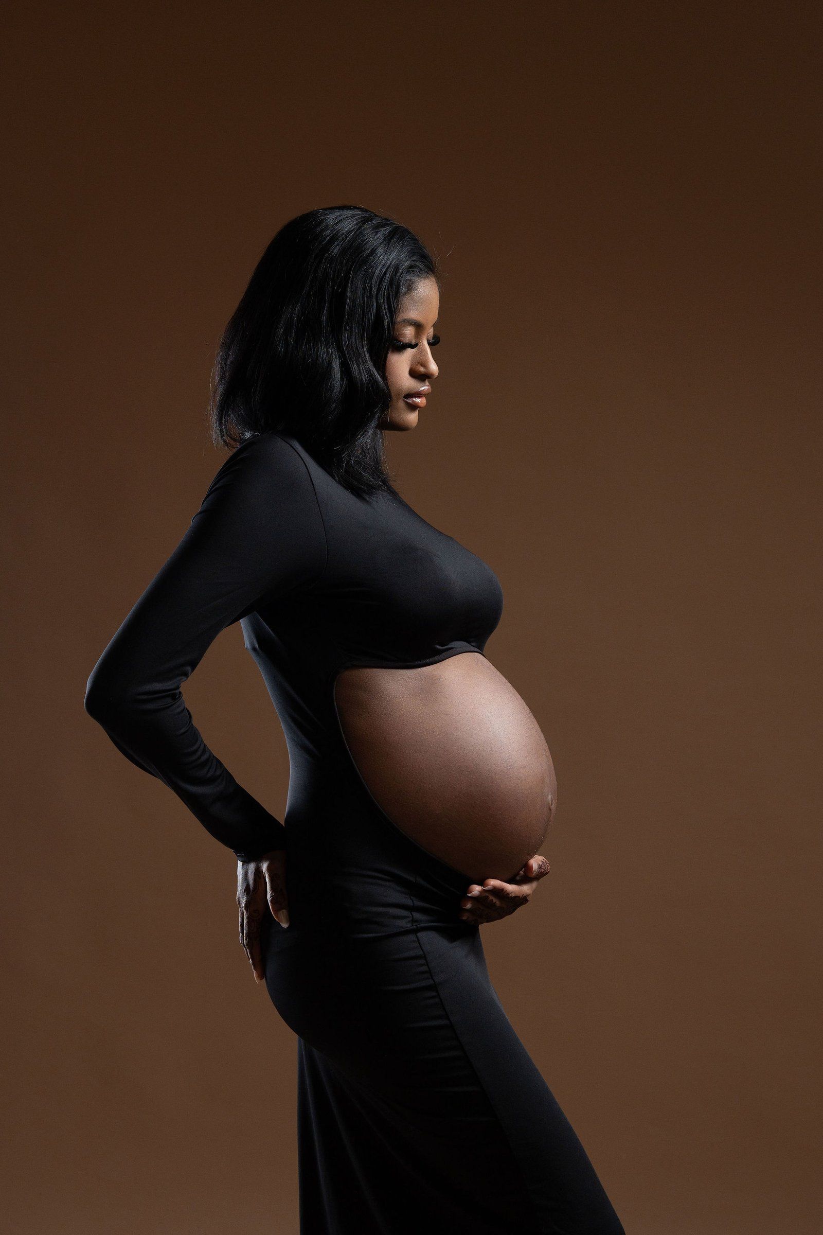 Pregnant Black woman in black dress cradling baby bump, studio portrait on brown background.