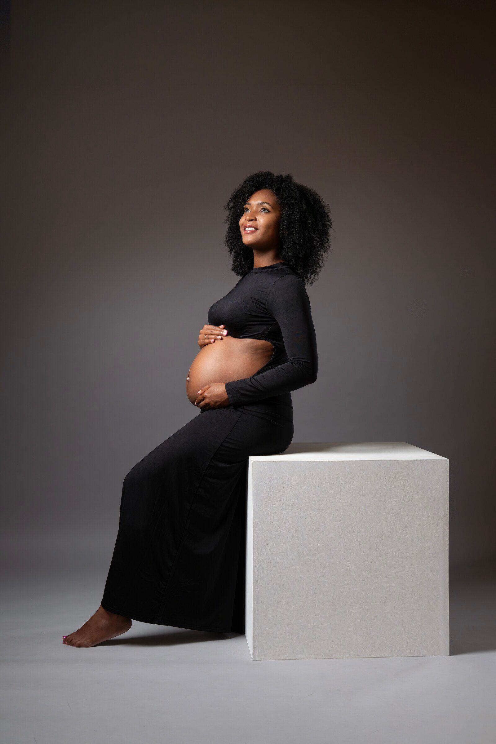 Smiling pregnant Black woman in black dress sits on white cube, cradling baby bump in studio portrait.