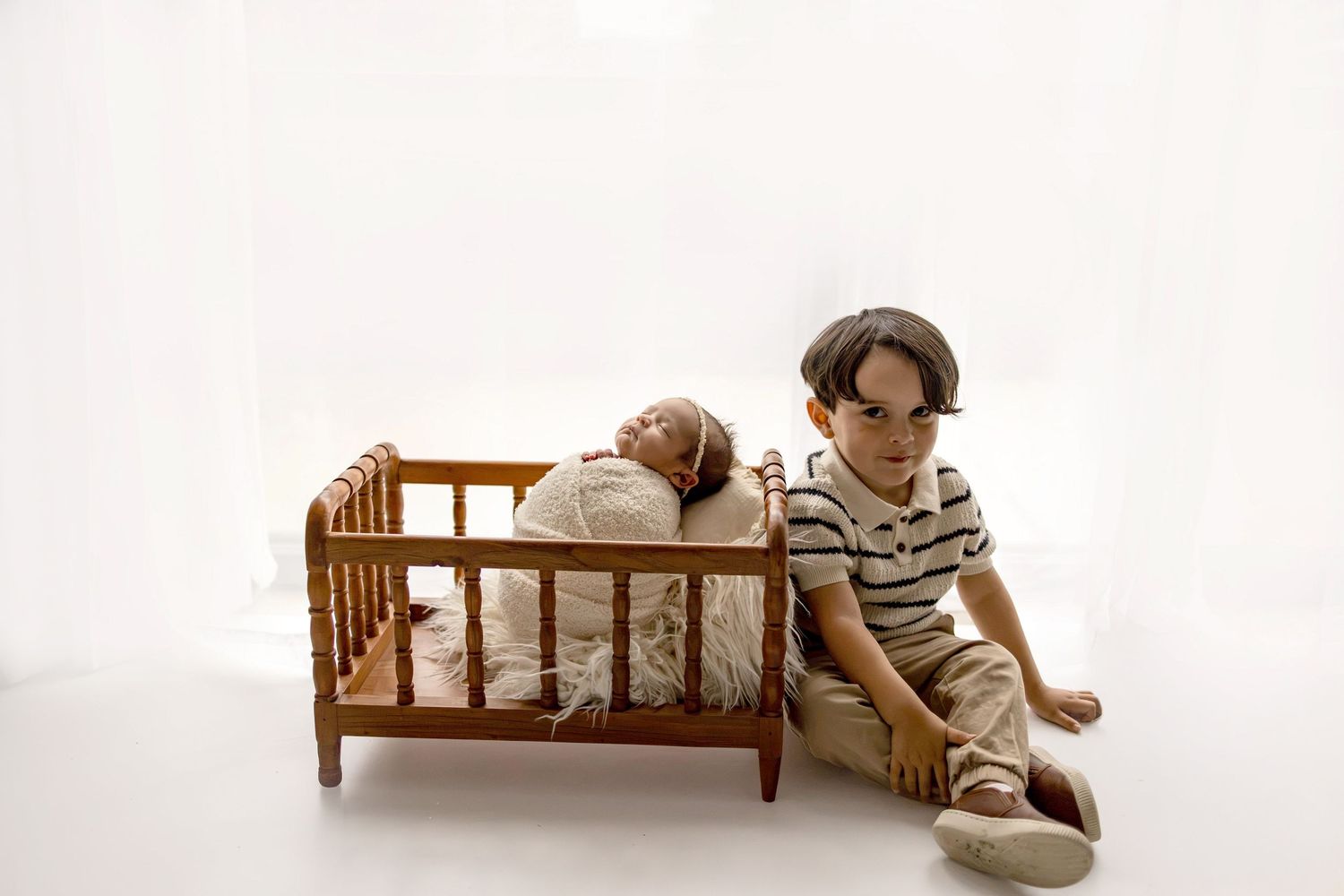 A young boy in a striped shirt sits beside a wooden toy crib with a sleeping newborn baby inside, on white background.