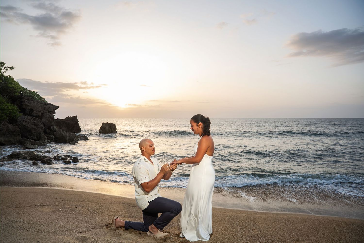 Man proposes to woman on sandy beach at sunset with ocean waves and golden sky in background.
