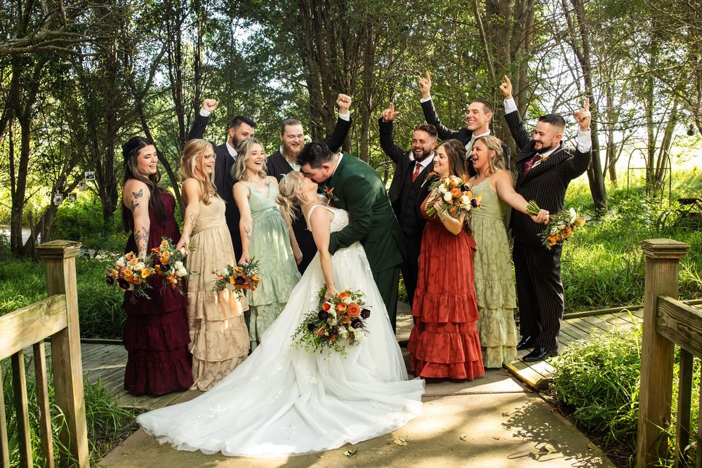 Wedding party celebrates as bride and groom kiss on a wooden bridge surrounded by lush green trees.