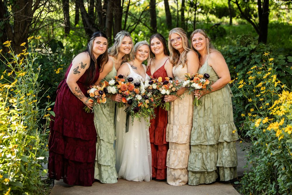 Bride in white gown poses with five bridesmaids in mismatched green and red dresses holding autumn bouquets outdoors.