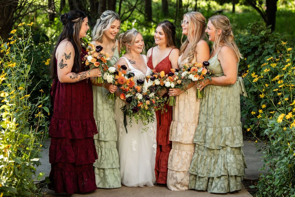 Bride in white gown with bridesmaids in green and burgundy dresses holding colorful bouquets outdoors.