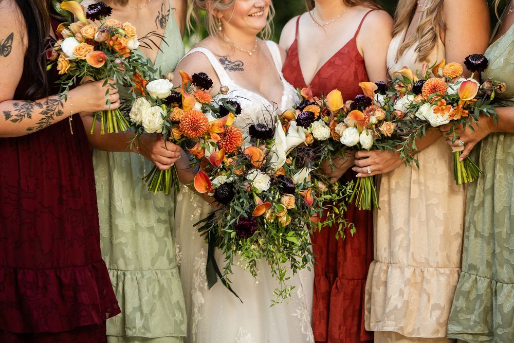 Bride and bridesmaids in mismatched burgundy and sage dresses holding lush fall bouquets with orange, red, and white blooms.