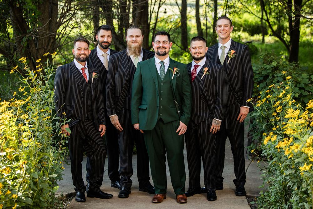 Groom in green suit poses with five groomsmen in dark suits and burgundy ties outdoors at a wedding.