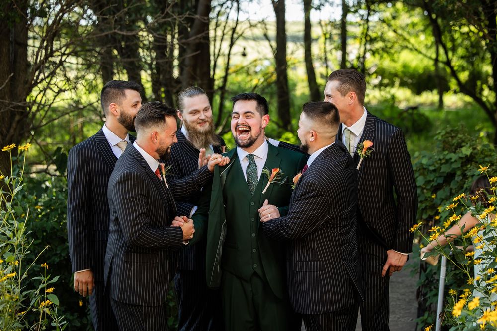 Groom in green suit laughing with groomsmen in pinstripe suits outdoors at a wedding celebration.