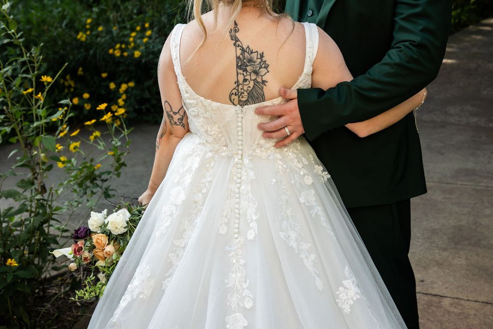 Bride in lace wedding gown with back tattoo and floral details, embraced by groom in green suit outdoors.