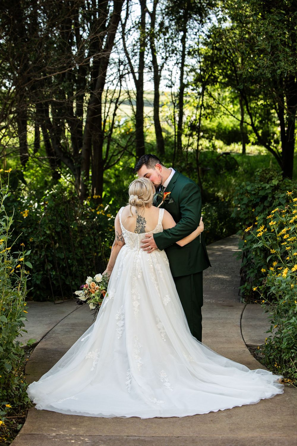 Bride and groom share a romantic kiss in a lush garden, bride wearing a lace gown with long train, groom in dark green suit.