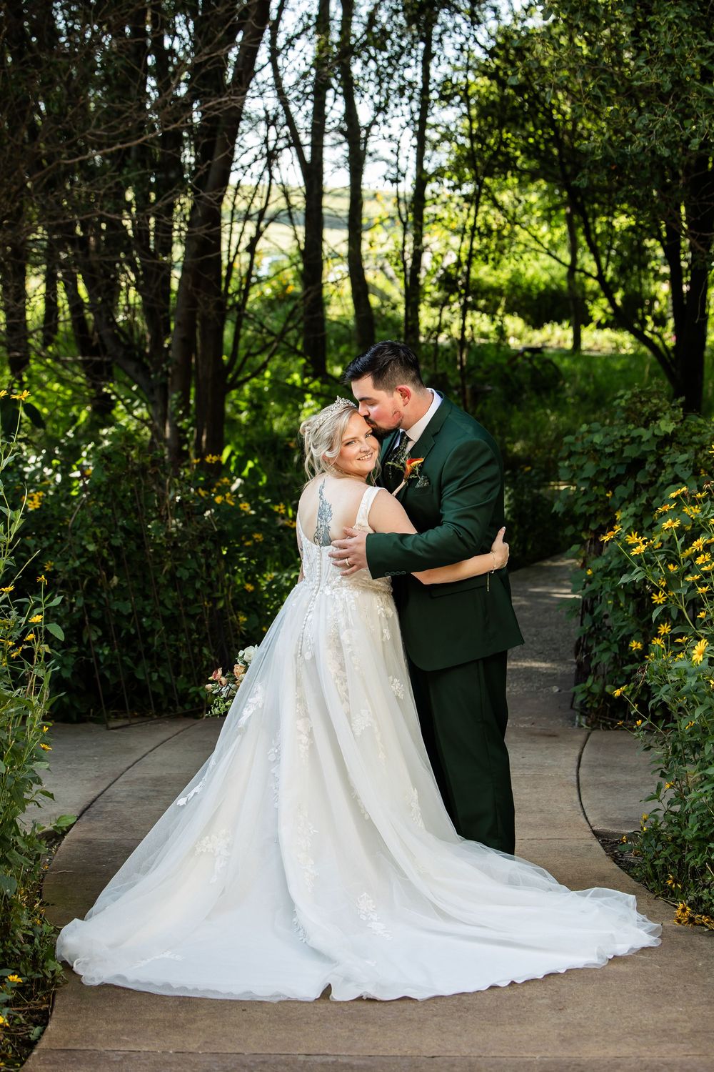 Bride and groom share a romantic kiss in a lush garden, bride in lace gown, groom in dark green suit.