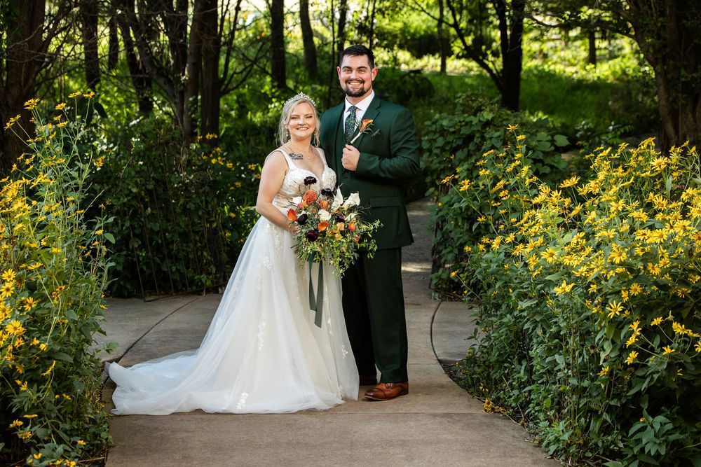 Bride and groom posing on a garden path surrounded by lush greenery and yellow flowers on their wedding day.