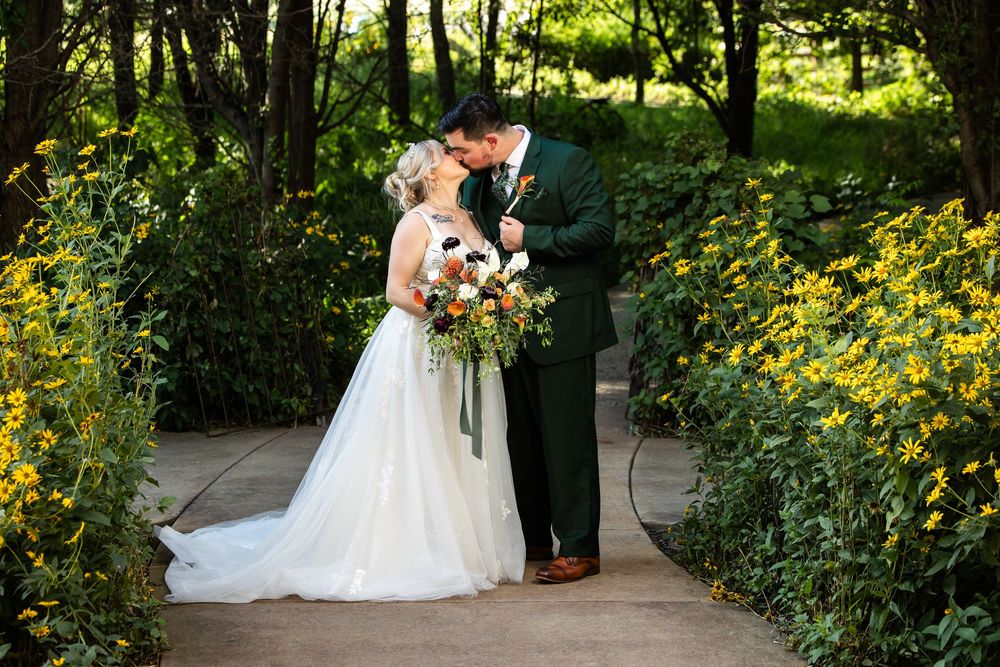 Bride and groom share a romantic kiss surrounded by lush greenery and yellow wildflowers on their wedding day.
