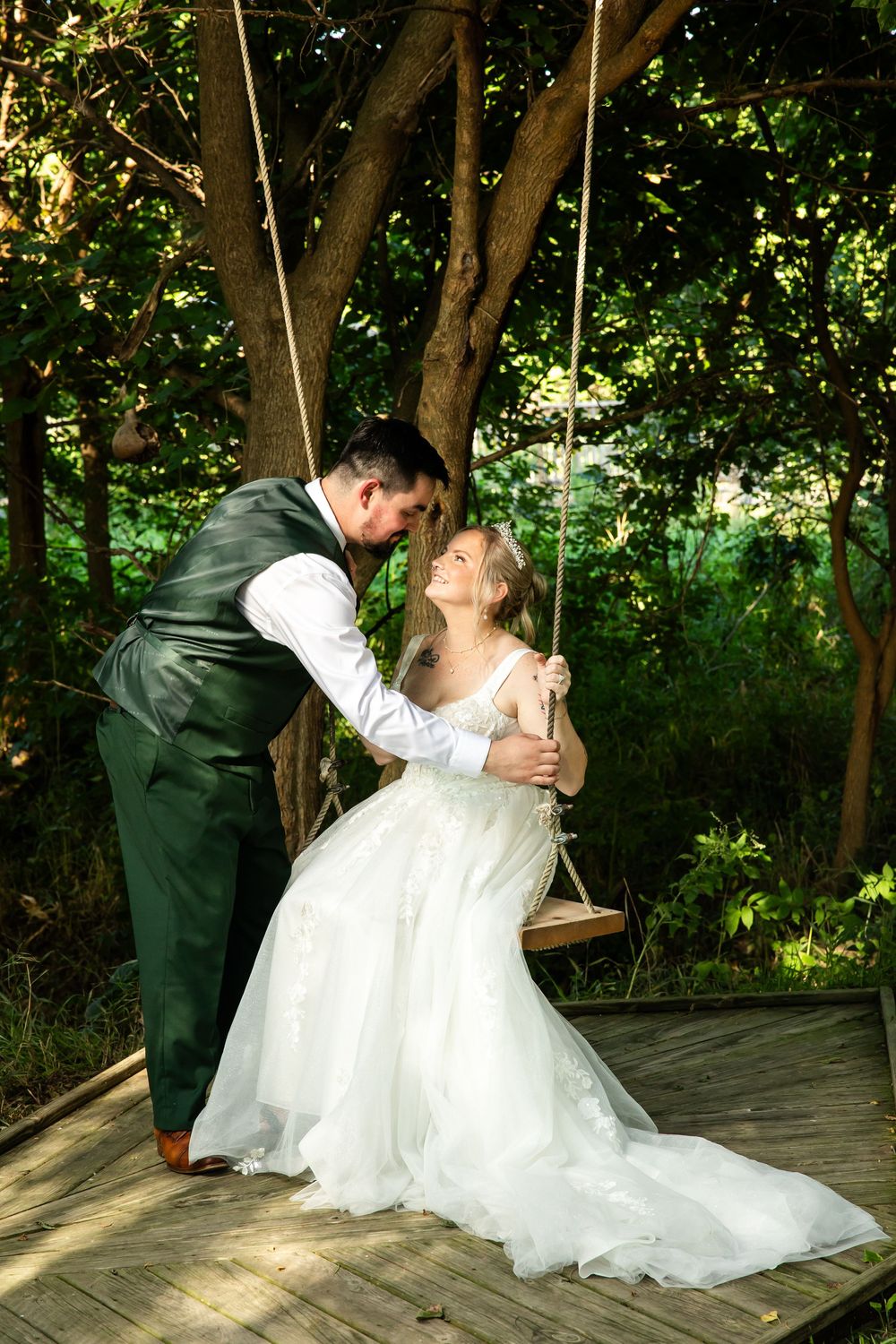Bride in white gown sits on wooden swing as groom in green suit leans in for a kiss in lush garden.