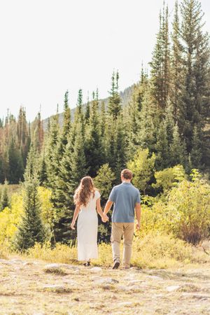 Couple holding hands walking toward pine forest in golden meadow, woman in white dress, man in blue shirt and khaki pants.