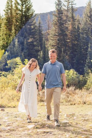 Couple holding hands walking through a mountain meadow, woman in white dress, man in blue polo shirt.