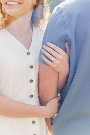 Close-up of smiling woman in white dress holding man's arm, showcasing a diamond engagement ring in soft sunlight.