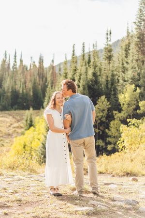 Couple embracing outdoors in a sunny mountain forest setting, woman in white dress, man in blue shirt and khaki pants.
