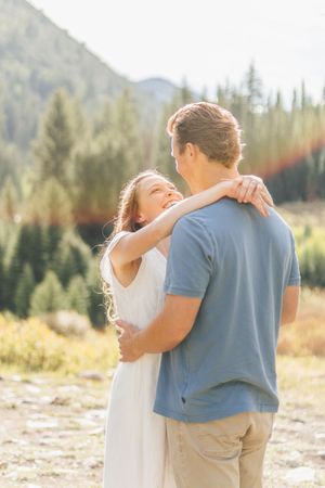 Couple embracing outdoors in mountain forest setting, woman in white dress smiling up at man in blue shirt.