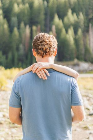 Man in blue t-shirt viewed from behind with arm around shoulder, standing before a lush green forest.