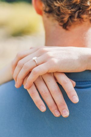 Close-up of a woman's hand with a diamond engagement ring resting on a man's shoulder.