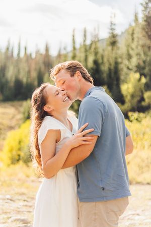 Couple embracing and sharing a kiss outdoors with pine trees and mountains in the background on a sunny day.