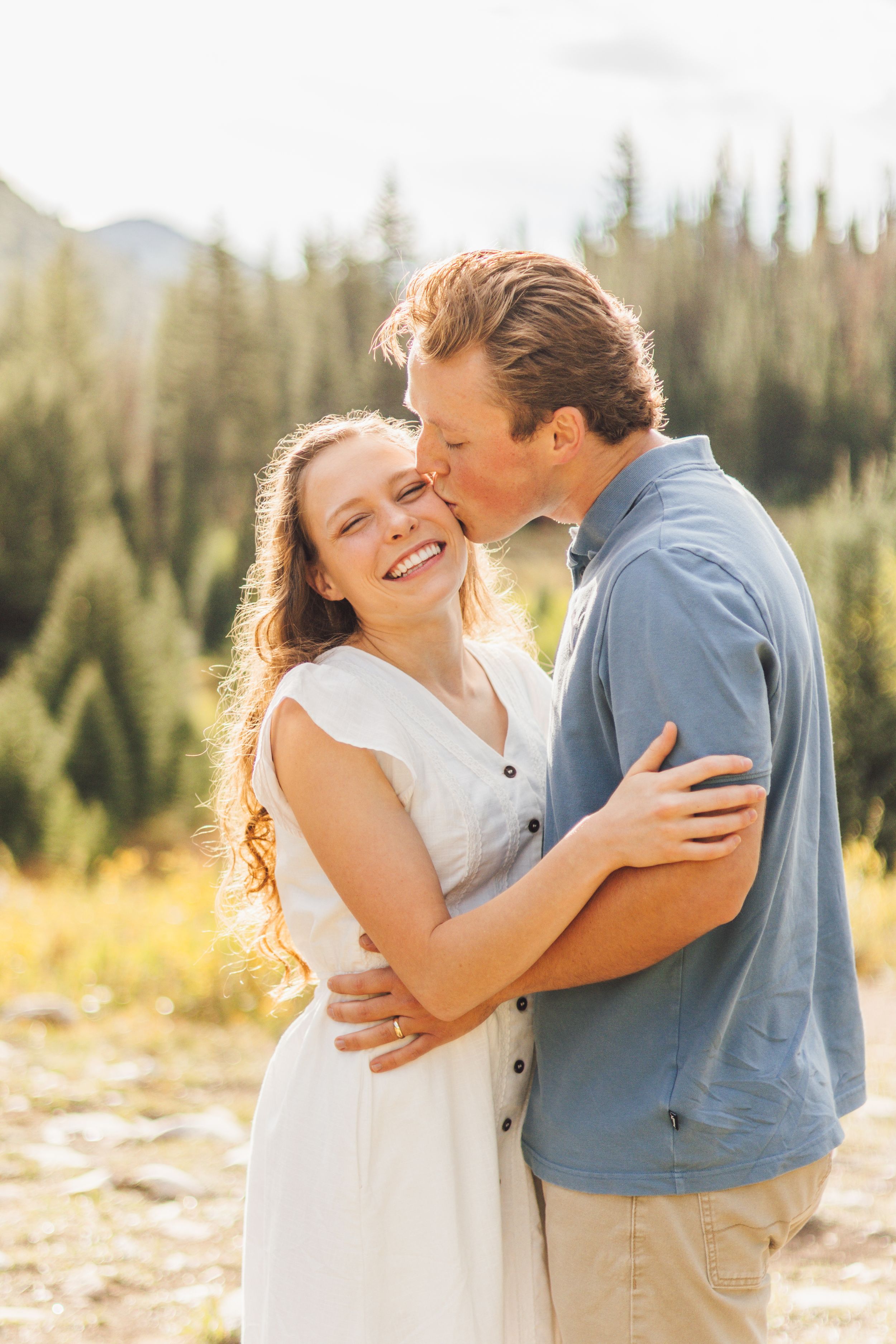 Couple embracing outdoors, man kissing woman on cheek, she smiles in white dress, pine forest background.