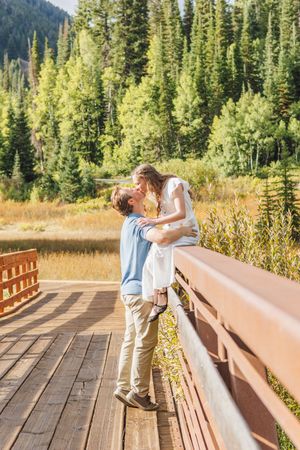 Couple sharing a romantic kiss on a wooden bridge surrounded by lush green pine trees and golden meadows.